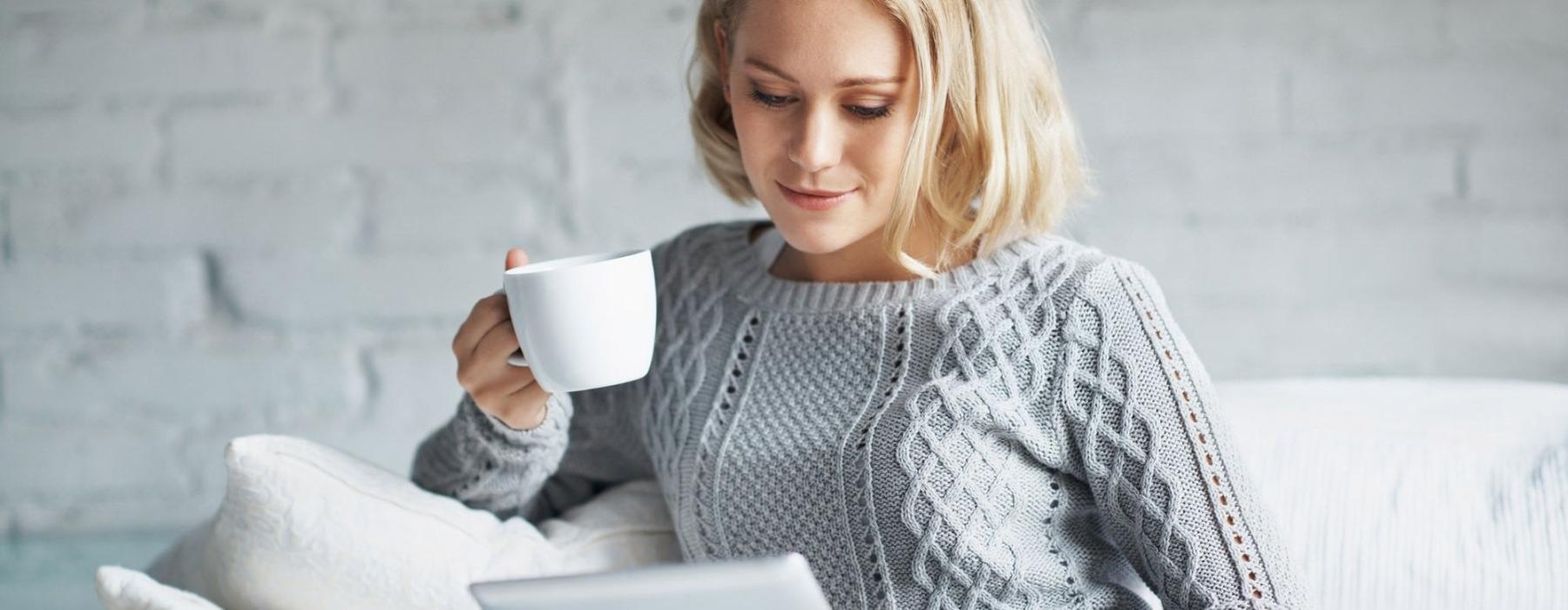 a woman sits on a couch with a cup of coffee and looks at her tablet